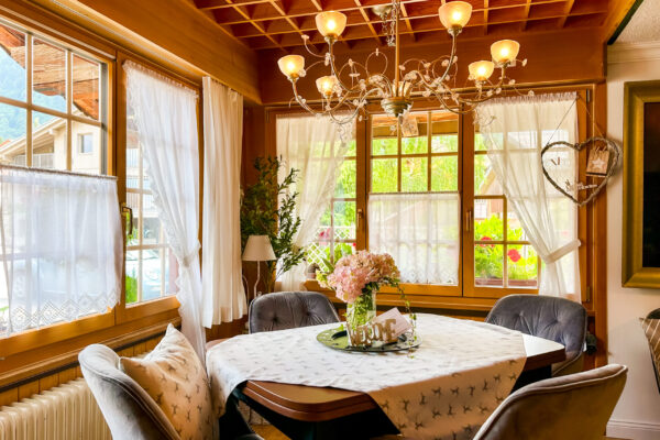 Chalet Simmi-39 Chalet Simmi: Sitzbereich in der Stube, wirkt sehr einladend mit warmen Licht. Ein Kronleuchter kommt aus der Holzdecke und darunter steht ein gedeckter Tisch mit Blumenstrauß und vier Stühlen drum herum, sehr edel eingerichtet